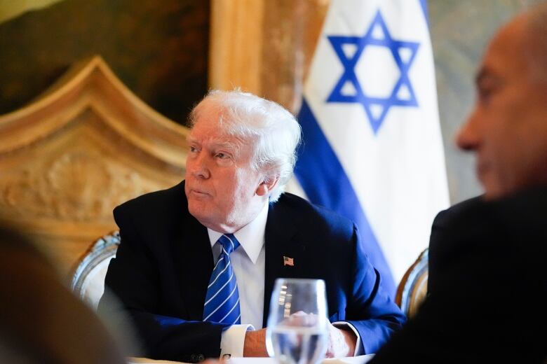 A man in a dark suit and blue tie sits at a table. The white-and-blue Israeli flag is behind him.