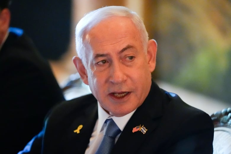 A man in a dark suit and blue tie is pictured. A pin with the American and Israeli flags is on his lapel.