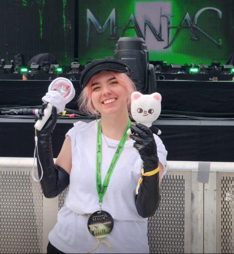 A woman with pink hair holds K-pop swag in front of a stage at a concert. A screen behind her reads 'Maniac'