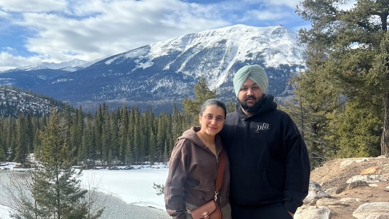 A man wearing a turban standing next to a woman outside with a snow-capped mountain, trees and water in the background. 