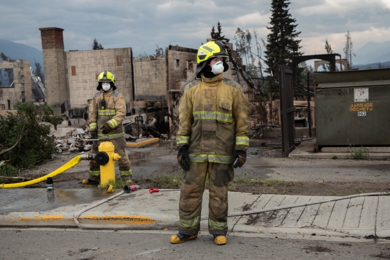 Firefighters in full gear stand before the shell of a burned out building. 