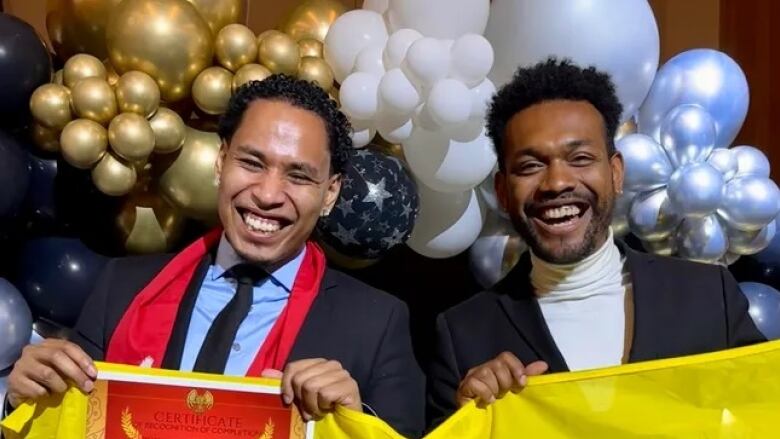 Two young Black men stand smiling in front of balloons, holding up a certificate and flag. The men are in suits. They are visible from the chest up.