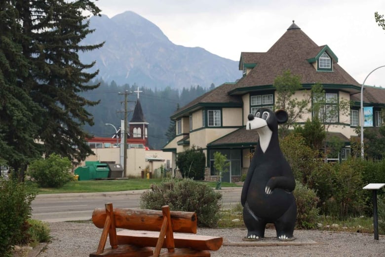 The black and white statue of Jasper the bear stands in a park in front of a historic beige building in Jasper. He has a rotund belly and thick legs and a white snout that pokes out with a black, round nose at the tip.