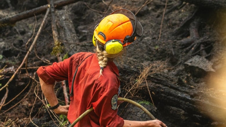 A person with a blonde braid holds a hose among charred forest