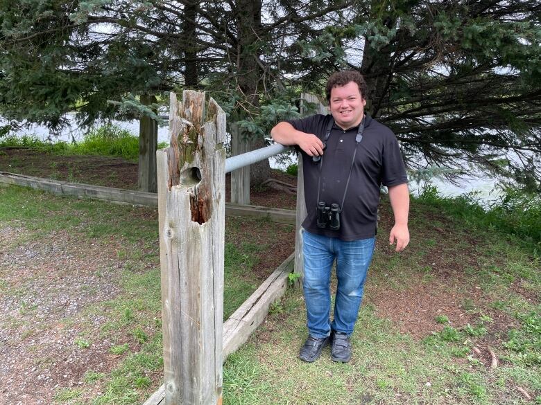 A young man leaning on a wooden fence with a pair of binoculars hanging from his neck.