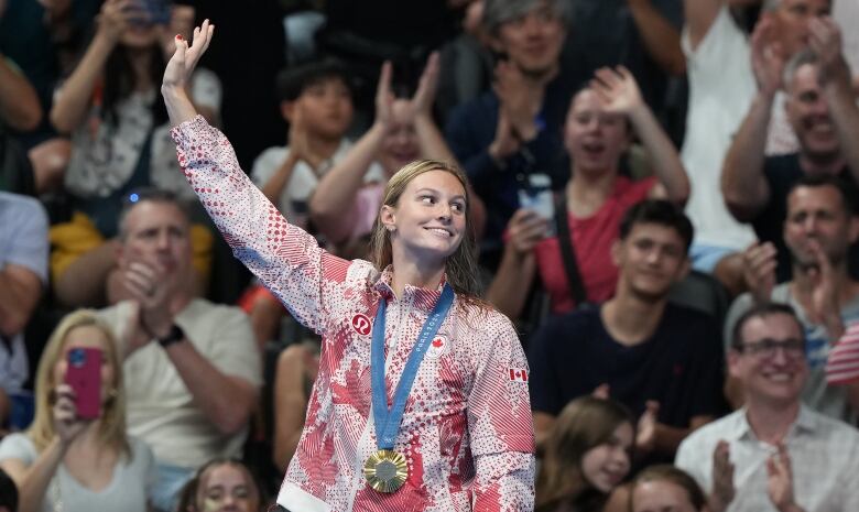 Female teen waving to a crowd wearing a gold medal around her neck following a swim competition.