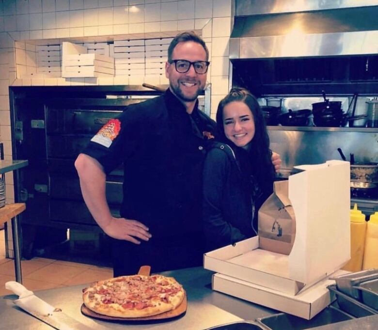 A man wearing glasses stands in a kitchen, smiling with his arm around a teenage girl.