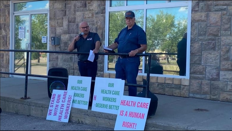 Two men speak at a rally, with signs beneath them reading 'Our Health Care Workers Deserve Better' and 'Health Care is a Human Right!'