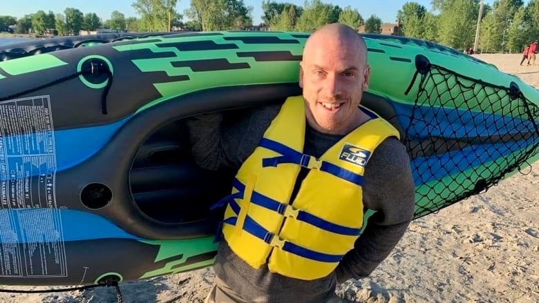 A man holds an inflatable boat on his back, wearing a life jacket and smiles at the camera.