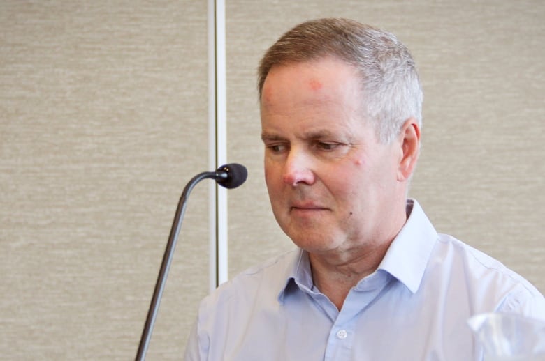 A man with short grey hair wearing a light blue shirt sits in front of a microphone.