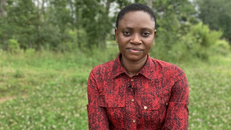 A woman sits on a park bench with grass and trees behind her. The woman is Black, and wearing a red button-up shirt.