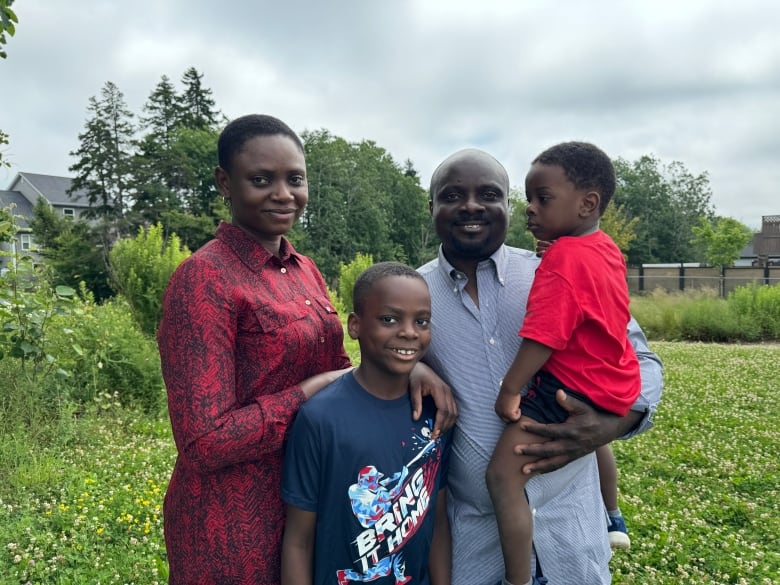 A family of four stand together for a photo standing on the grass in a park. The family are a mother, father, eight year old boy, and three year old boy in his father's arms. 