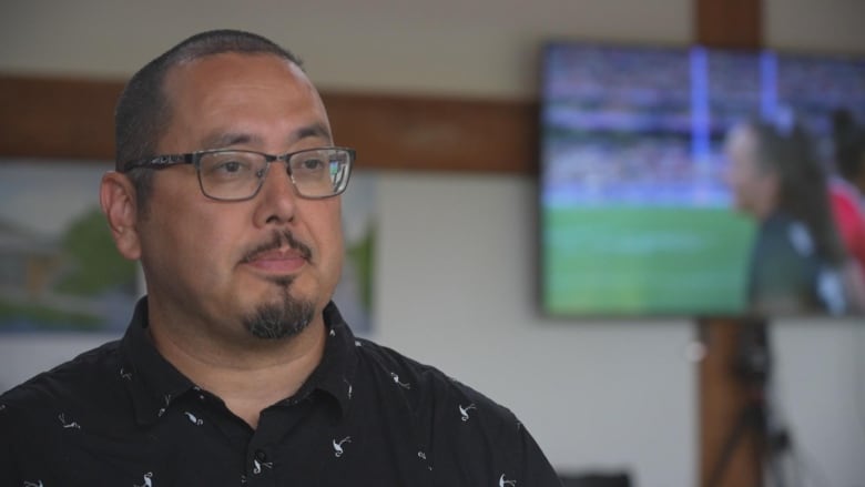 A man wearing glasses and a TV screen showing rugby sevens in the background