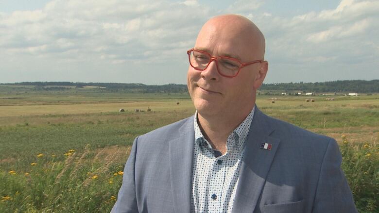 A bald man with red-framed glasses and wearing a grey suit stands in front of a grassy field.