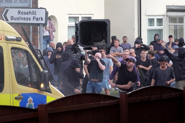 A man throws a large garbage can in the direction of a police van.