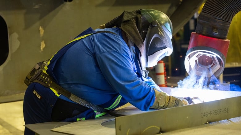 A man in a welding mask and blue coveralls makes the ceremonial first steel cut to begin construction of the CCGS Sermilik Glacier Arctic and Offshore Patrol Vessel. 