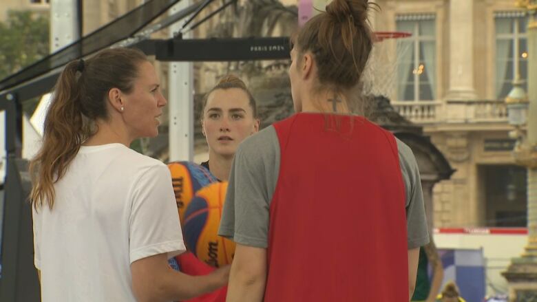 Three women in basketball practice uniforms huddle together on the court, talking about strategy. The coach, wearing right, is holding to blue-and-yellow basketballs.