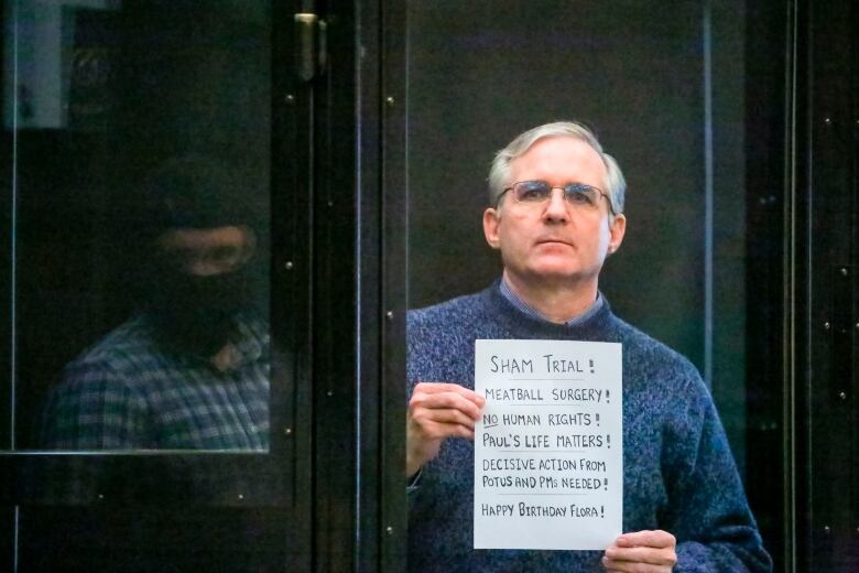 A bespectacled clean shaven man wearing a collared shirt underneath a sweater holds up a handmade sign with written messages while standing behind a glass partition in what appears to be a kind of prisoner's box.