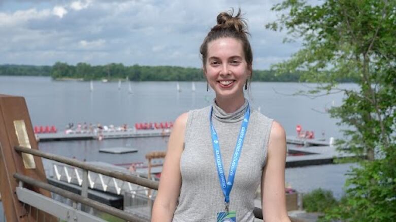 Elizabeth Grater, a woman, stands centre frame, smiling, before the backdrop of the Ottawa River at the NCC River House.