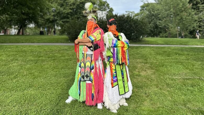 Two people dressed in colourful, ankle-length clothing stand together in a grassy park.