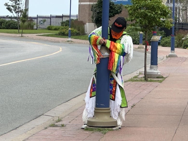 A person wearing a black baseball cap and colourful, ankle-length clothing ties a red cloth around a blue street lamp. 