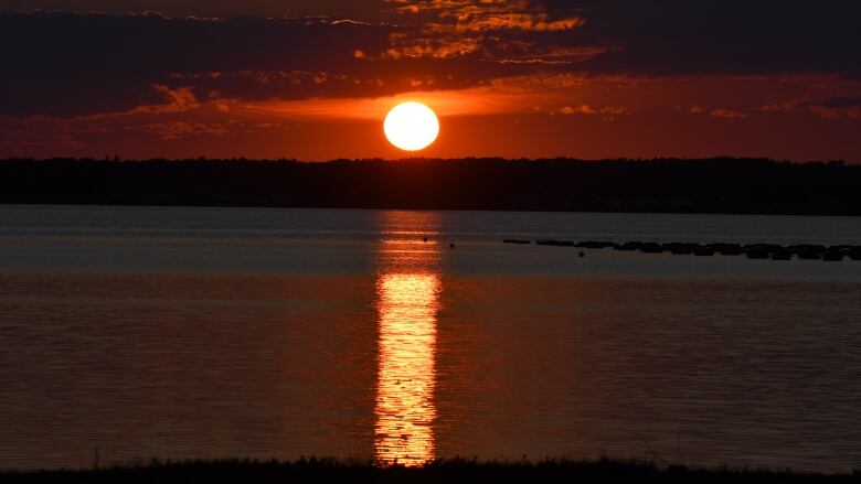 A blazing red sun hovers just over a tree line, its glow reflecting on a lake and turning the sky a deep orange.