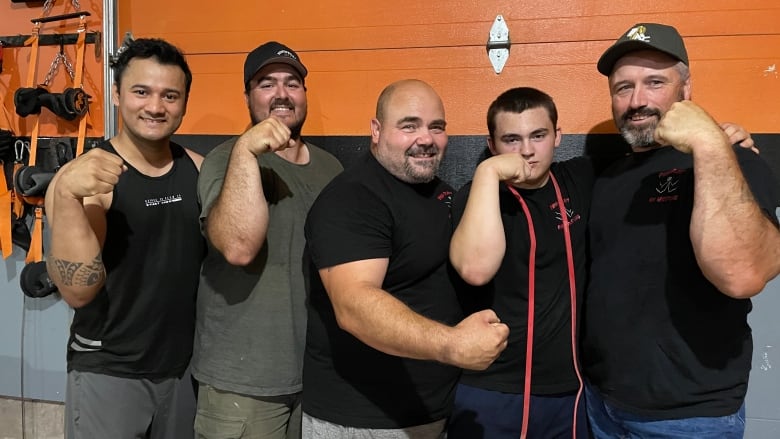 Five guys stand side by side in front of an orange and black wall. They are all smiling and flexing their arm muscle. 