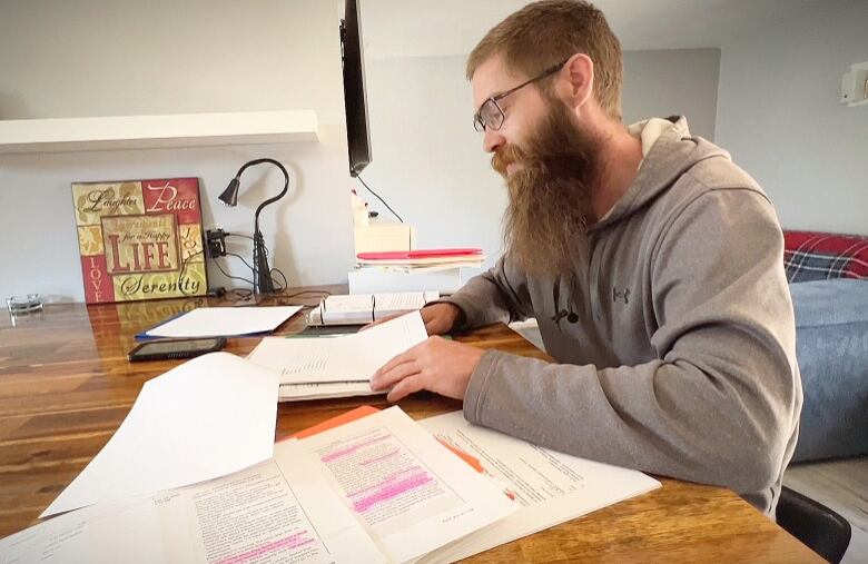 A man with short reddish hair and a long red beard wearing a grey hoodie and glasses looks over a stack of papers.