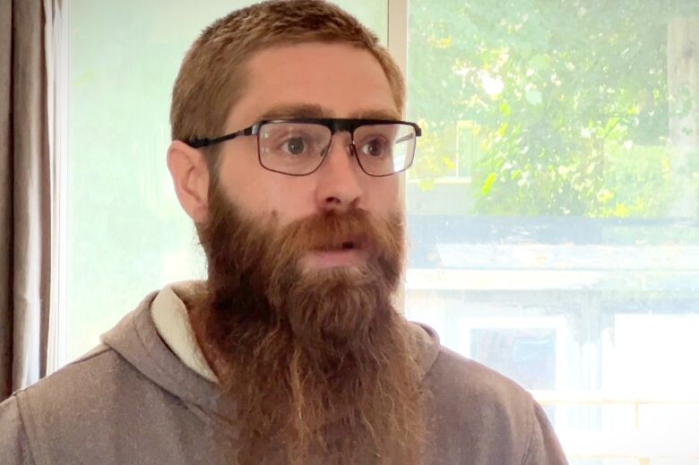 A man with short reddish hair and a long red beard wearing a grey hoodie and glasses speaks in front of a window.