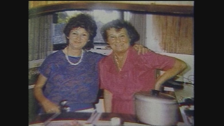 Two women dressed in vintage-looking clothing stand in a kitchen. 