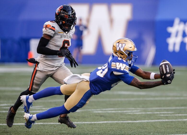 A football player in a blue and gold uniform catches a football while a player on the opposing team runs behind him. 