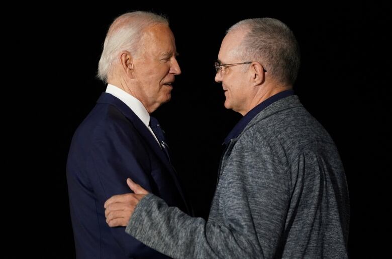 An older man in a blue suit greats another man in a grey sweater.