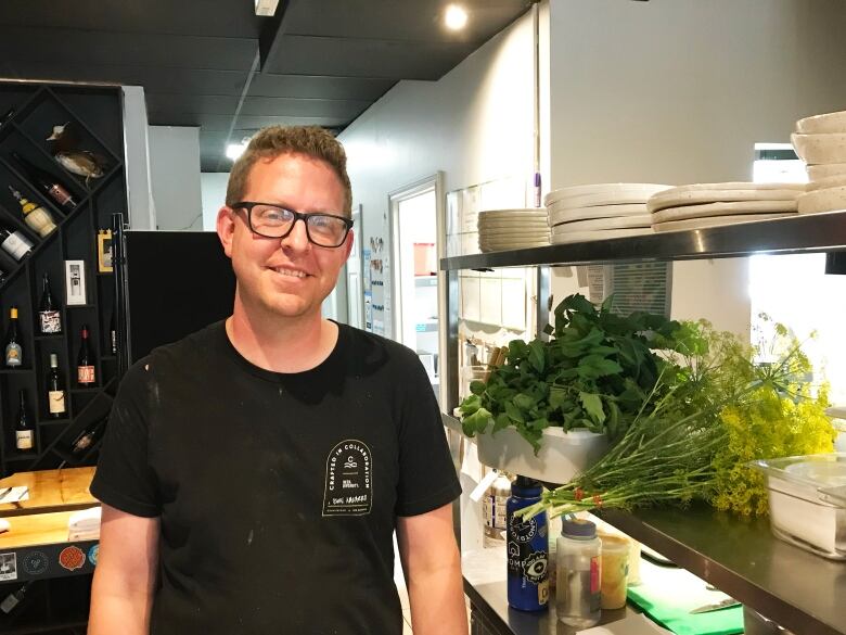 A chef stands in his kitchen. He is wearing a black shirt.