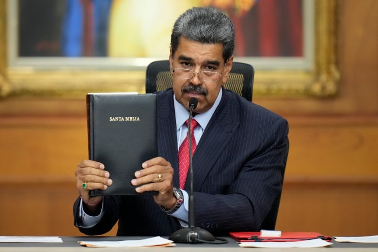 A mustachioed man in a suit and tie holds up a bible while sitting at a desk.