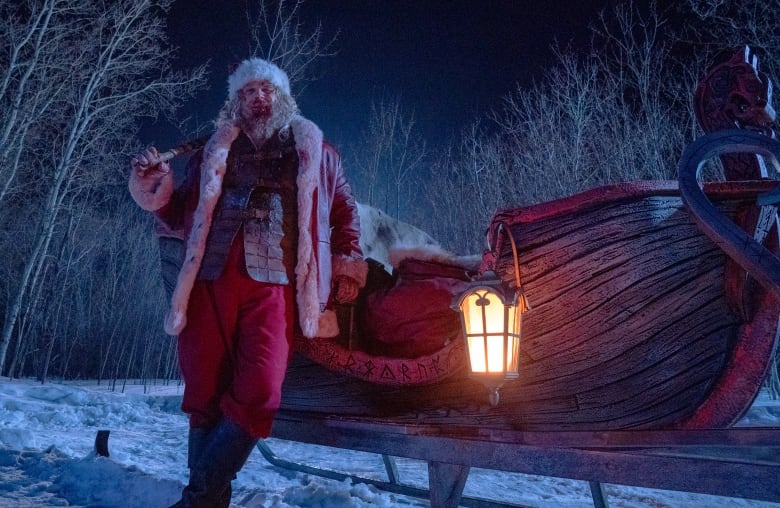 A bloodied man in a Santa Claus suit stands next to a wooden sleigh.