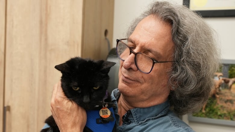 A man with long grey hair and glasses holds a black cat in his arms.