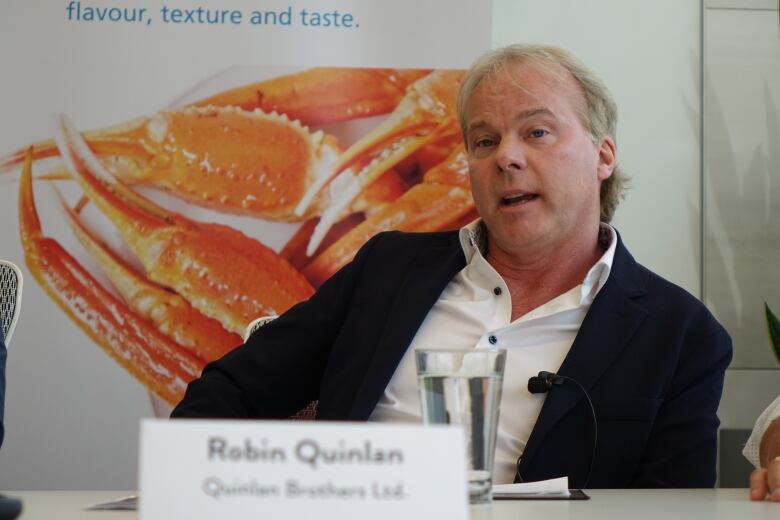 A man speaks to reporters at a table with a picture of a crab in the background