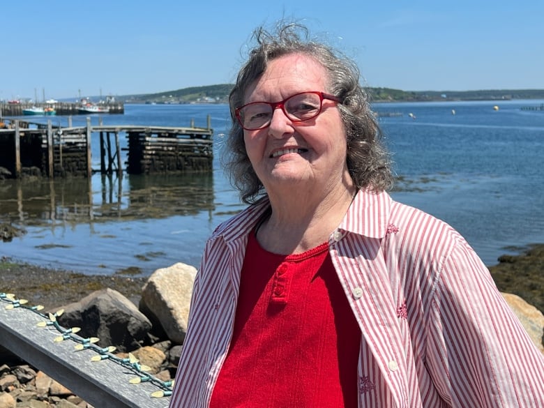 A woman in a red shirt and red glasses smiles. In the background is a wharf and ocean.