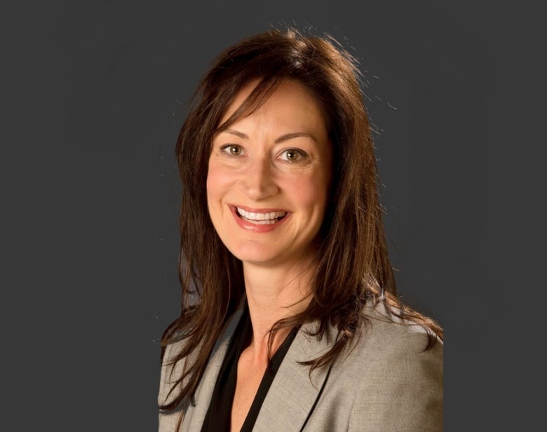 A woman with long brown hair and a light grey suit jacket smiles in front of a dark background.