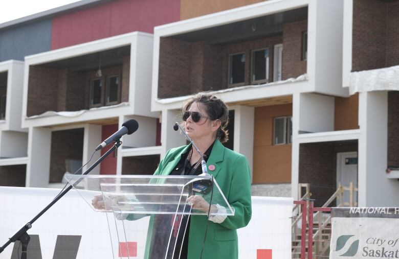 A woman with dark hair and wearing a green blazer speaks at a podium with construction behind her.