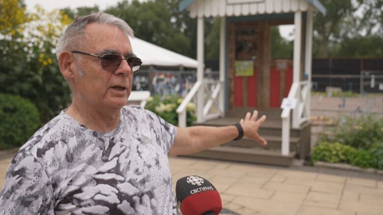 A middle aged man in a t-shirt and sunglasses motions to a patch of ground where the Island clubhouse once stood. Trees and parkland surround it. A cafe is seen beside it. It's daytime