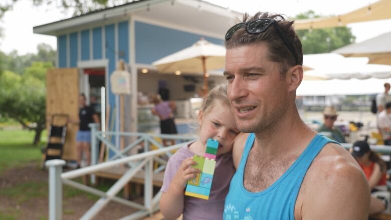 A man in a tank top is seen from chest up outside an outdoor cafe, holding a little girl in his arms. It's a grey day