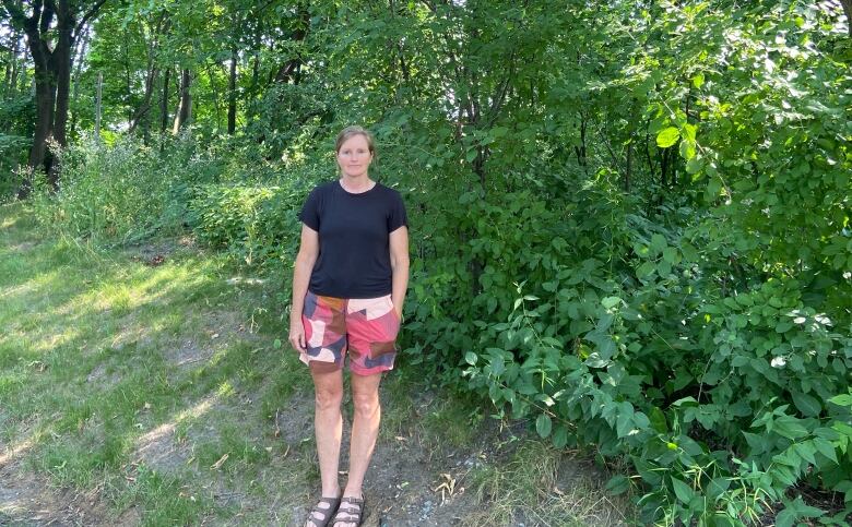 A woman wearing a black t-shirt and multicoloured shorts stands against a backdrop of trees and buckthorn in Atlantis Woods.