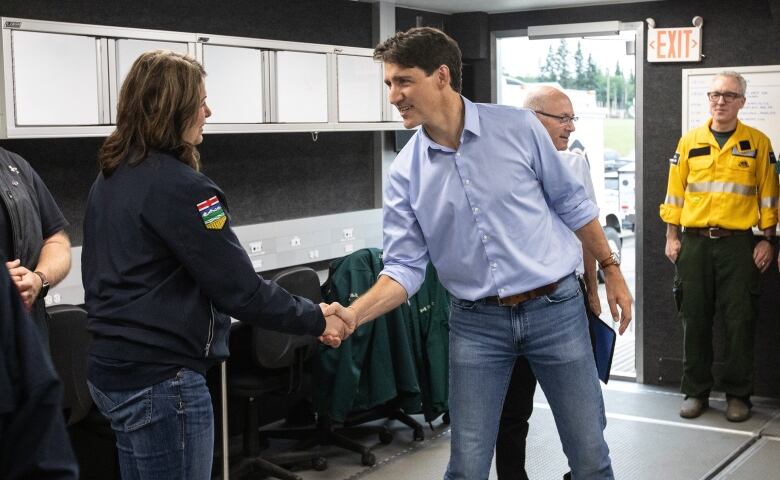 Prime Minister Justin Trudeau meets with Alberta Premier Dannielle Smith. They are shaking hands and saying something to one another. Trudeau is in a blue button up and blue jeans, while Smith is wearing a black jacket and blue jeans. They are in a small room with dark walls and fluorescent lighting. 