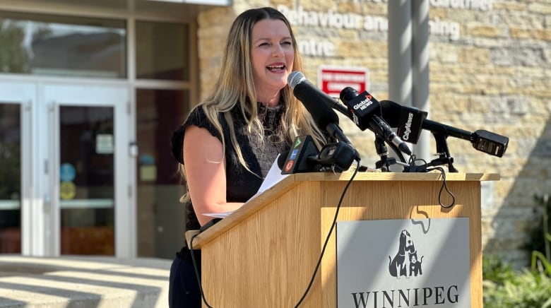 A woman with medium-length, dirty blond hair and wearing a black dress speaks at a podium.