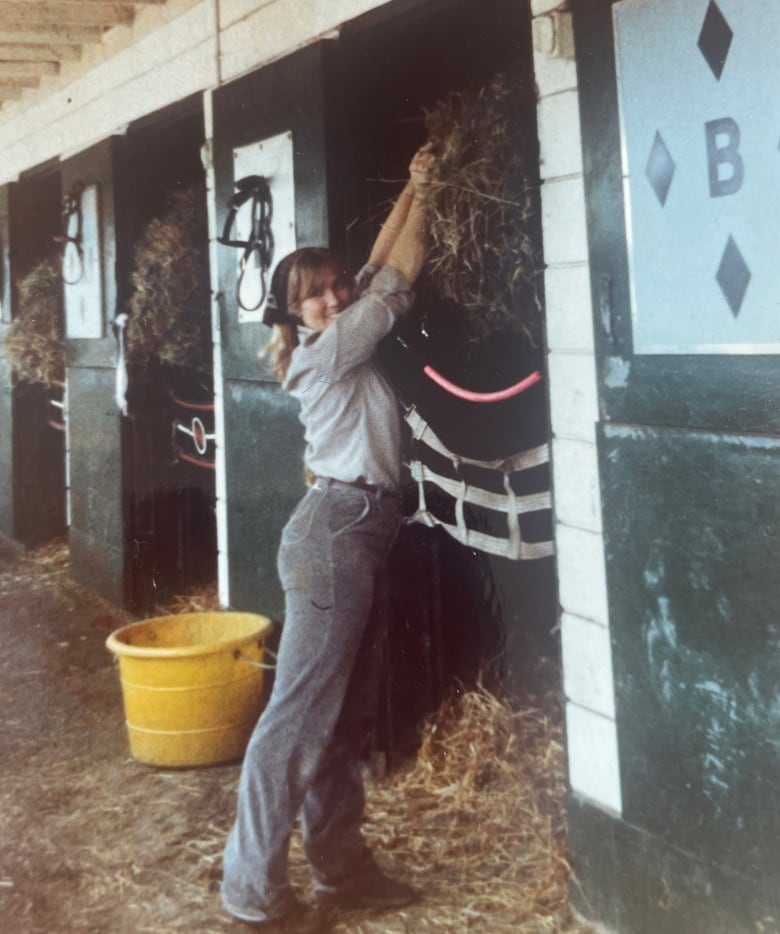 A woman turns to face the camera while loading a bushel of hay into a stable. 