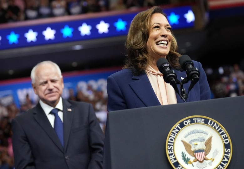 A woman in a navy blazer smiles and speaks into a microphone as a man waits behind her. 