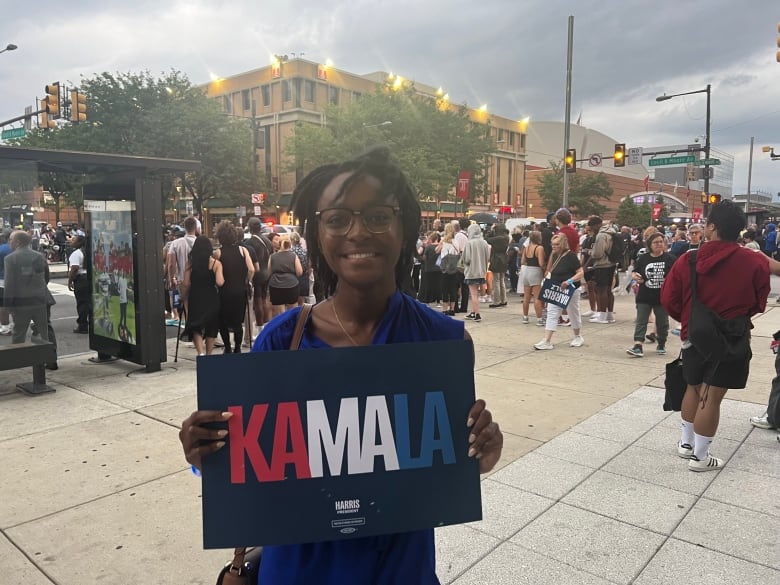 An African American girl wearing a blue dress is holding a sign that says KA-Ma-LA in red white and blue. There are dozens of people on the street behind her.