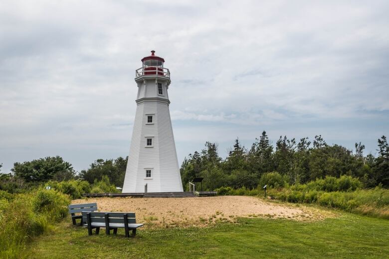 A white lighthouse sits in front of trees and behind two benches.