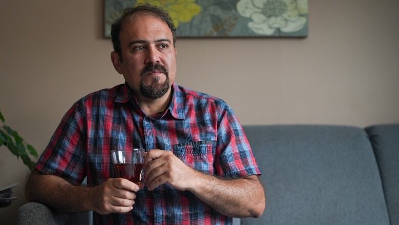 man with checkered shirt sits on couch holding tea